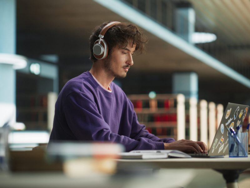 Thoughtful College Student Using Laptop Computer to Study in a Modern Library. Handsome Smart Man Learning Online, Getting Ready for Semester Exams, Drafting an Essay for Economics
